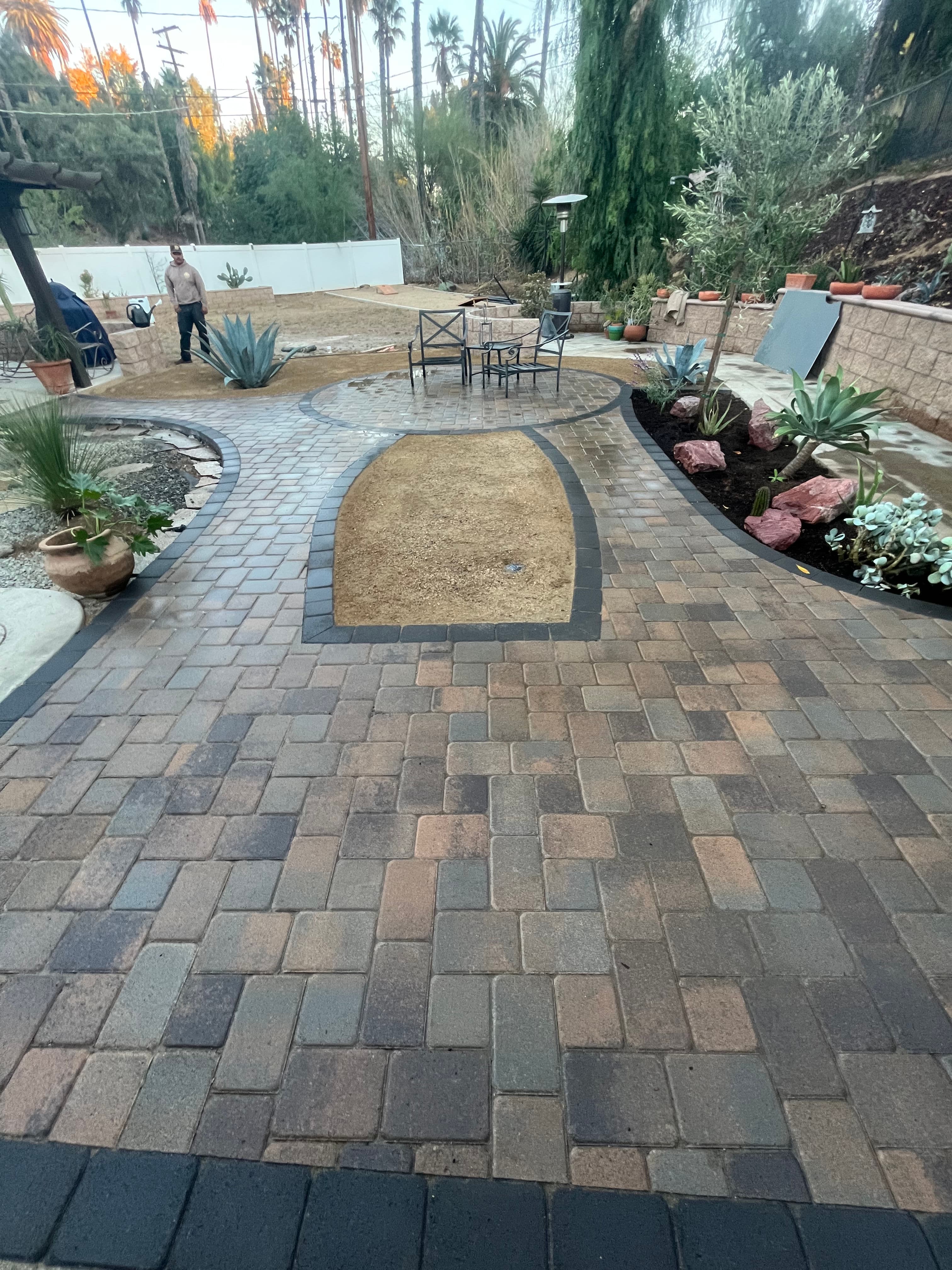 Backyard landscape with paving stones, planting areas, and a person working in greenery.